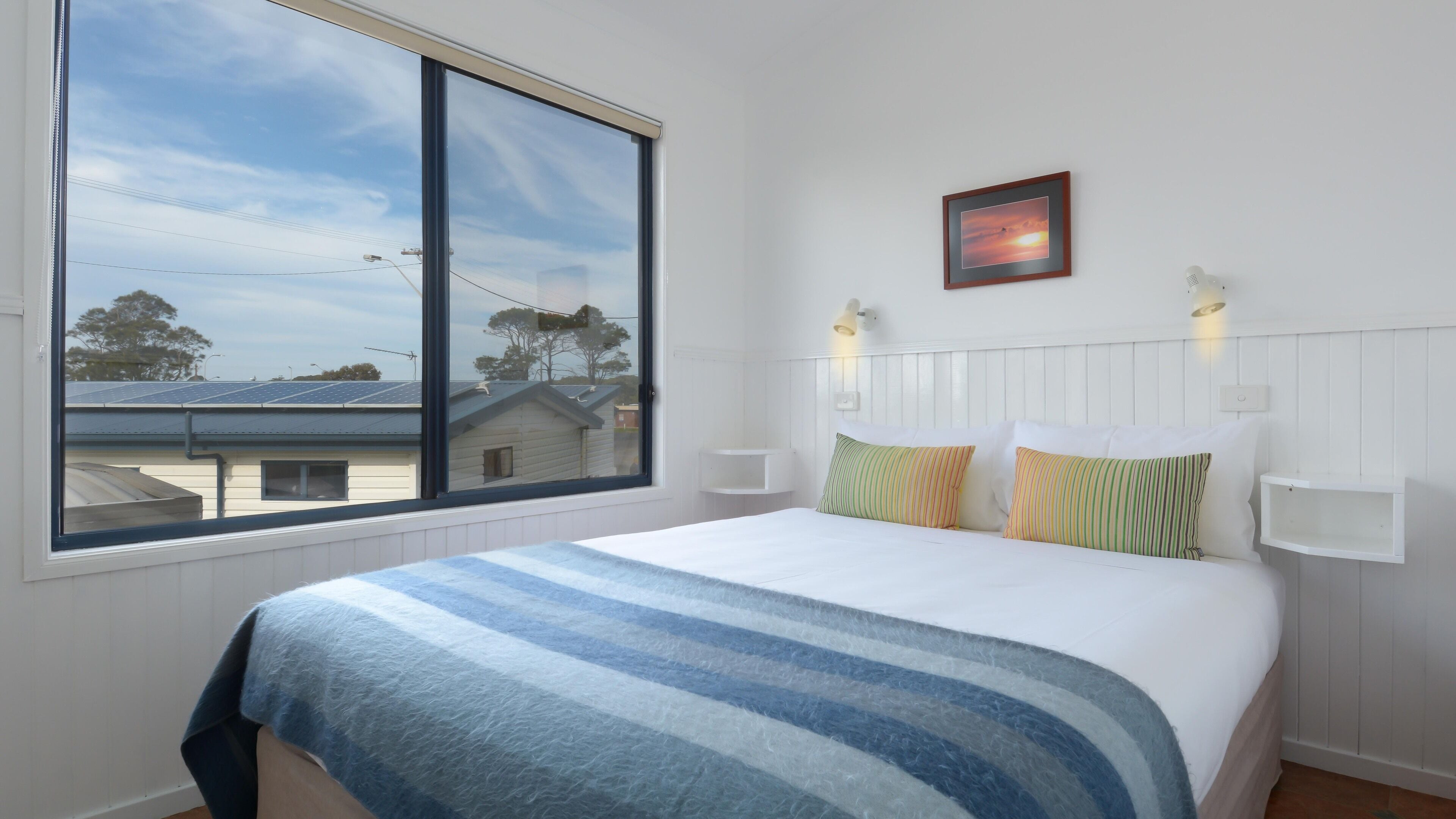 A bedroom with a large window, a bed with striped bedding, and two wall-mounted lamps.