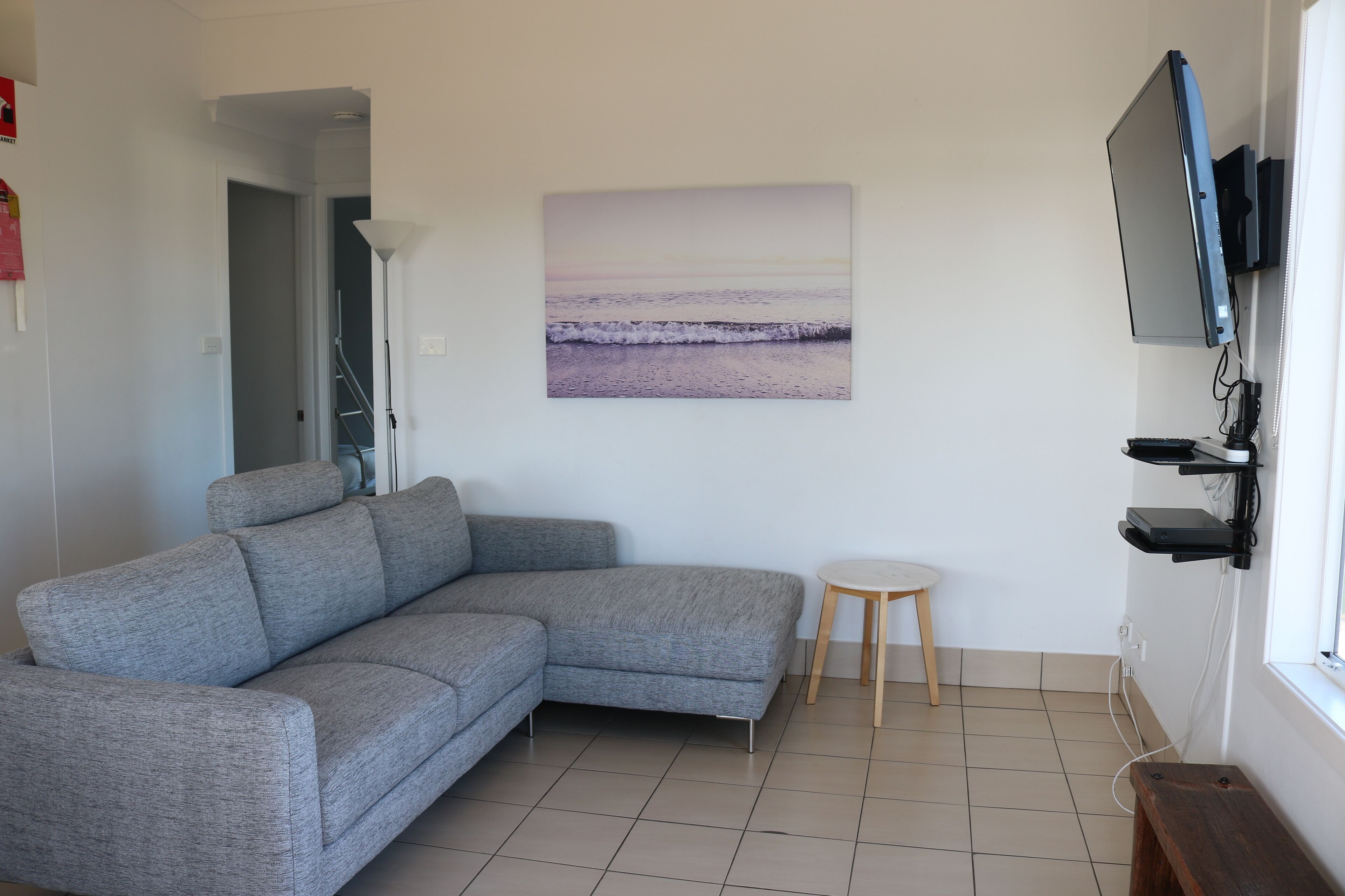 Een moderne woonkamer met een grijze modulaire bank, een klein rond tafeltje, een schilderij van een strandgezicht en een wandgemonteerde televisie.