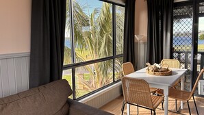 Una habitación con un ventanal grande, una mesa blanca con sillas de mimbre, un sofá marrón y vistas a palmeras y una masa de agua.