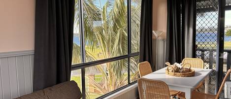 A room with a large window, a white table with wicker chairs, a brown sofa, and a view of palm trees and a body of water.