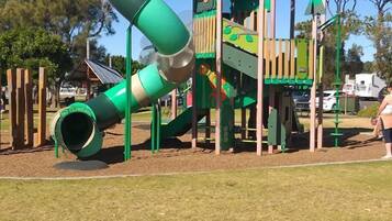 Explore the playground's spiral slide, climbing wall, and tower with a window, surrounded by lush greenery and outdoor seating areas.
