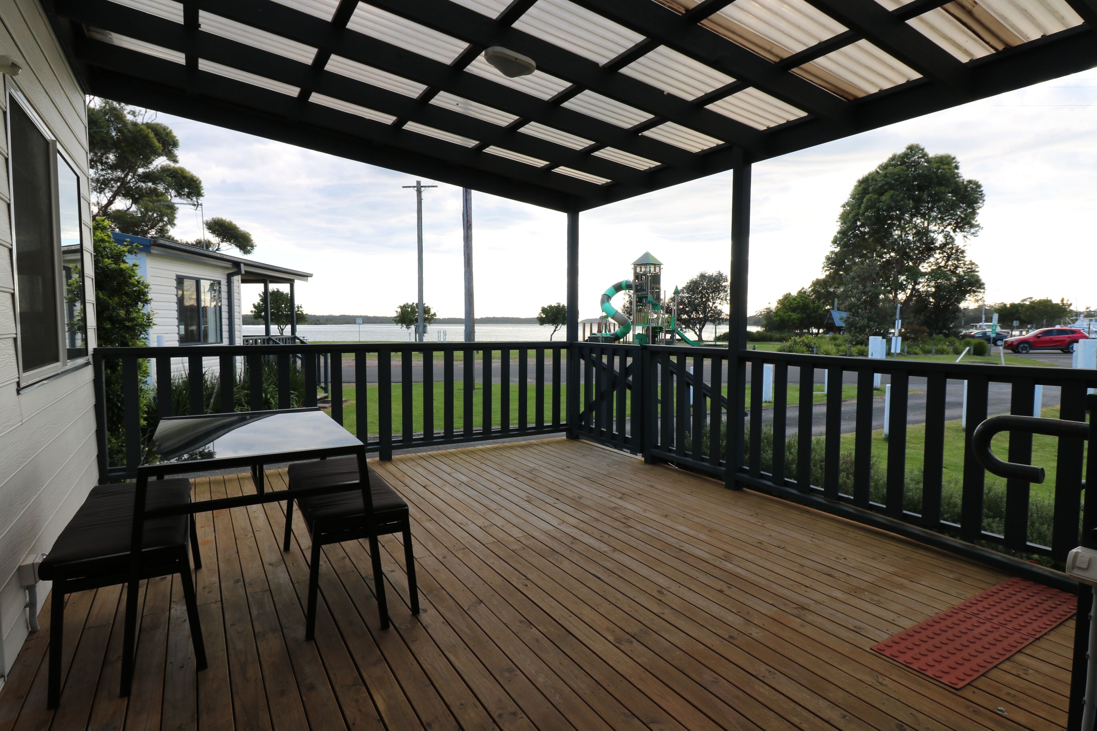 A wooden deck with a table and chairs, overlooking a water body and a playground.