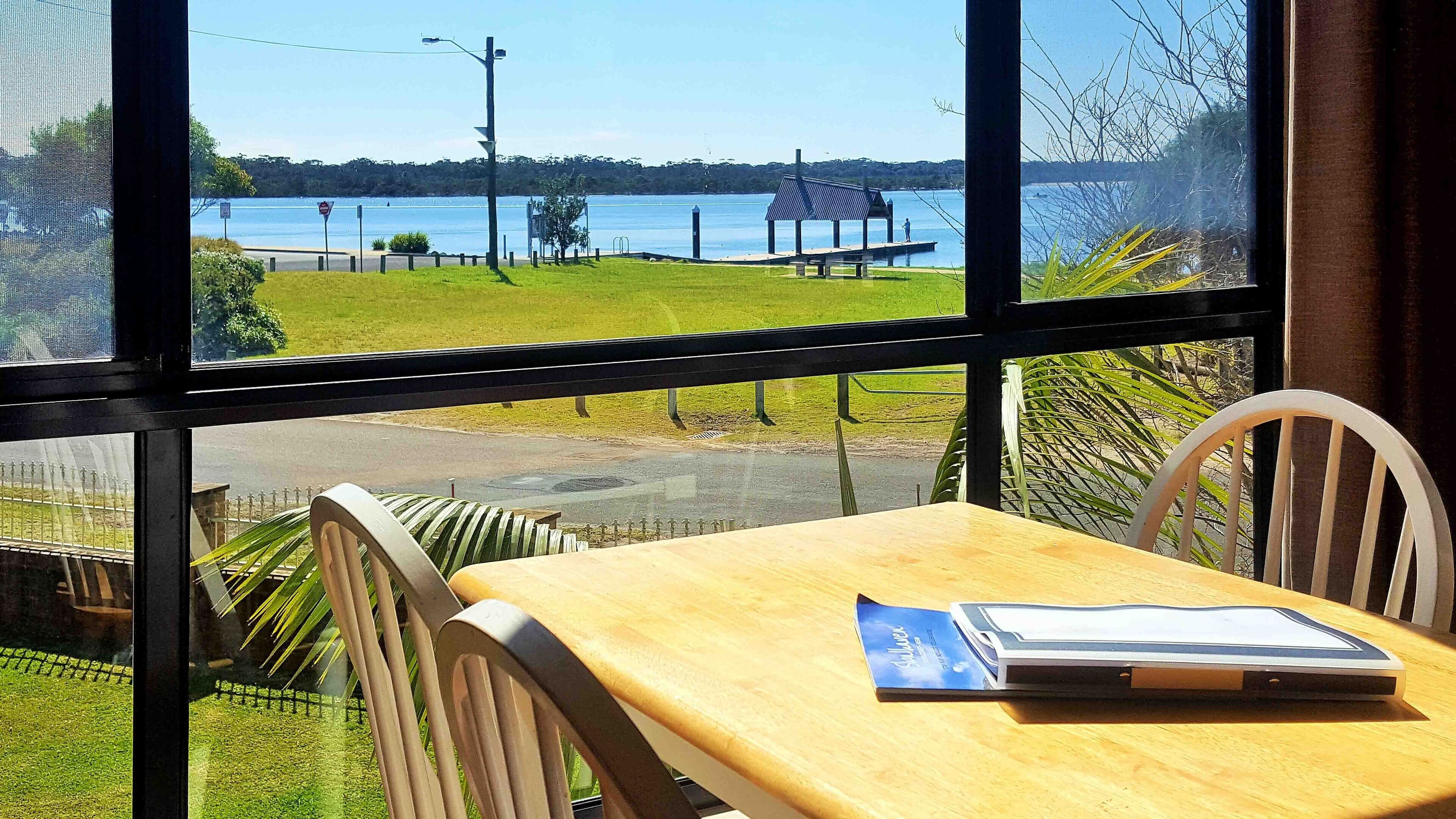 A room with a large window offering a view of a lake, a grassy area, and a dock. There is a wooden table with a chair and a book on it.