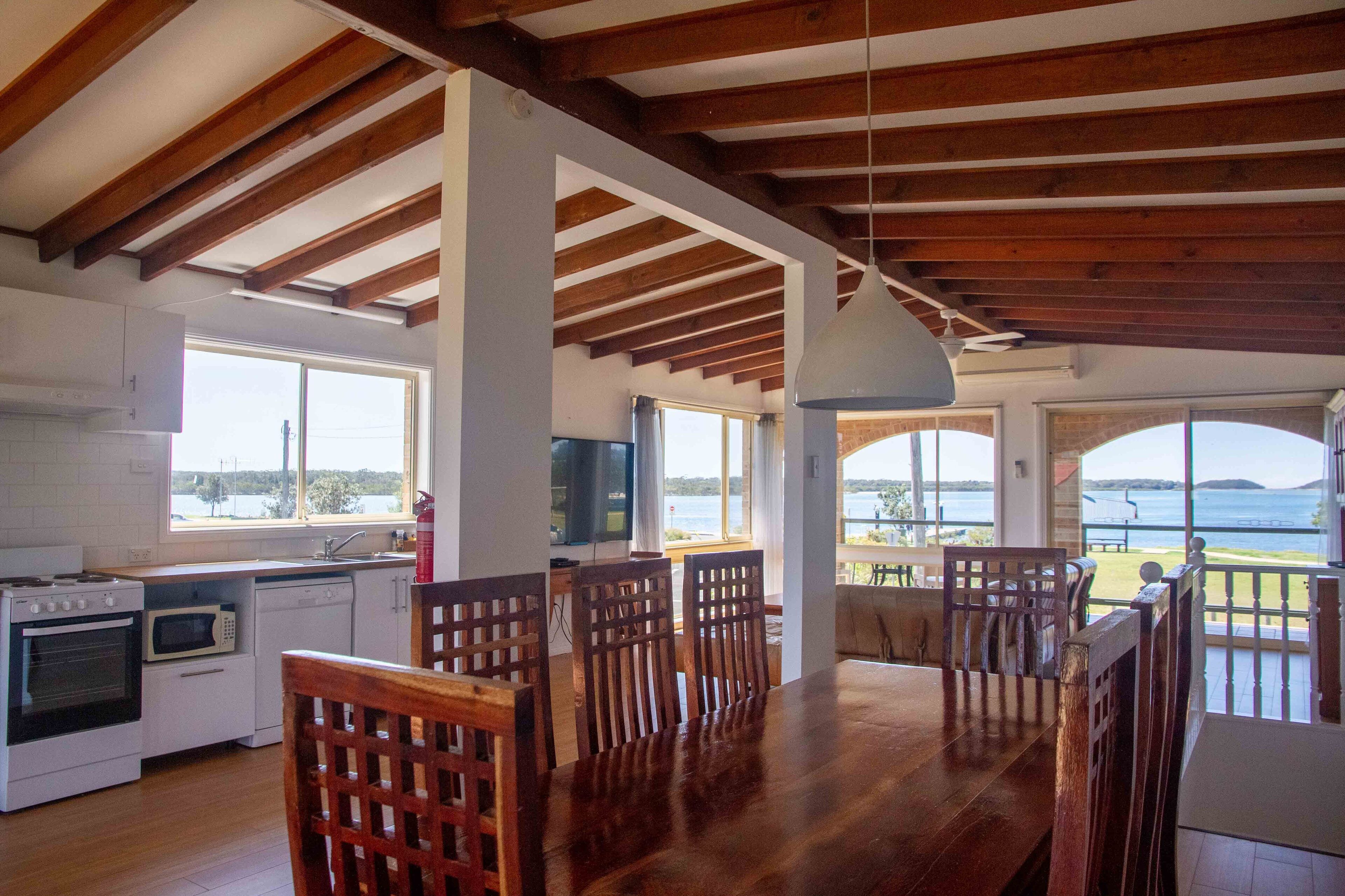 Una cocina amplia con vigas de madera, un comedor con una mesa grande y sillas, y vista al mar a través de ventanales.