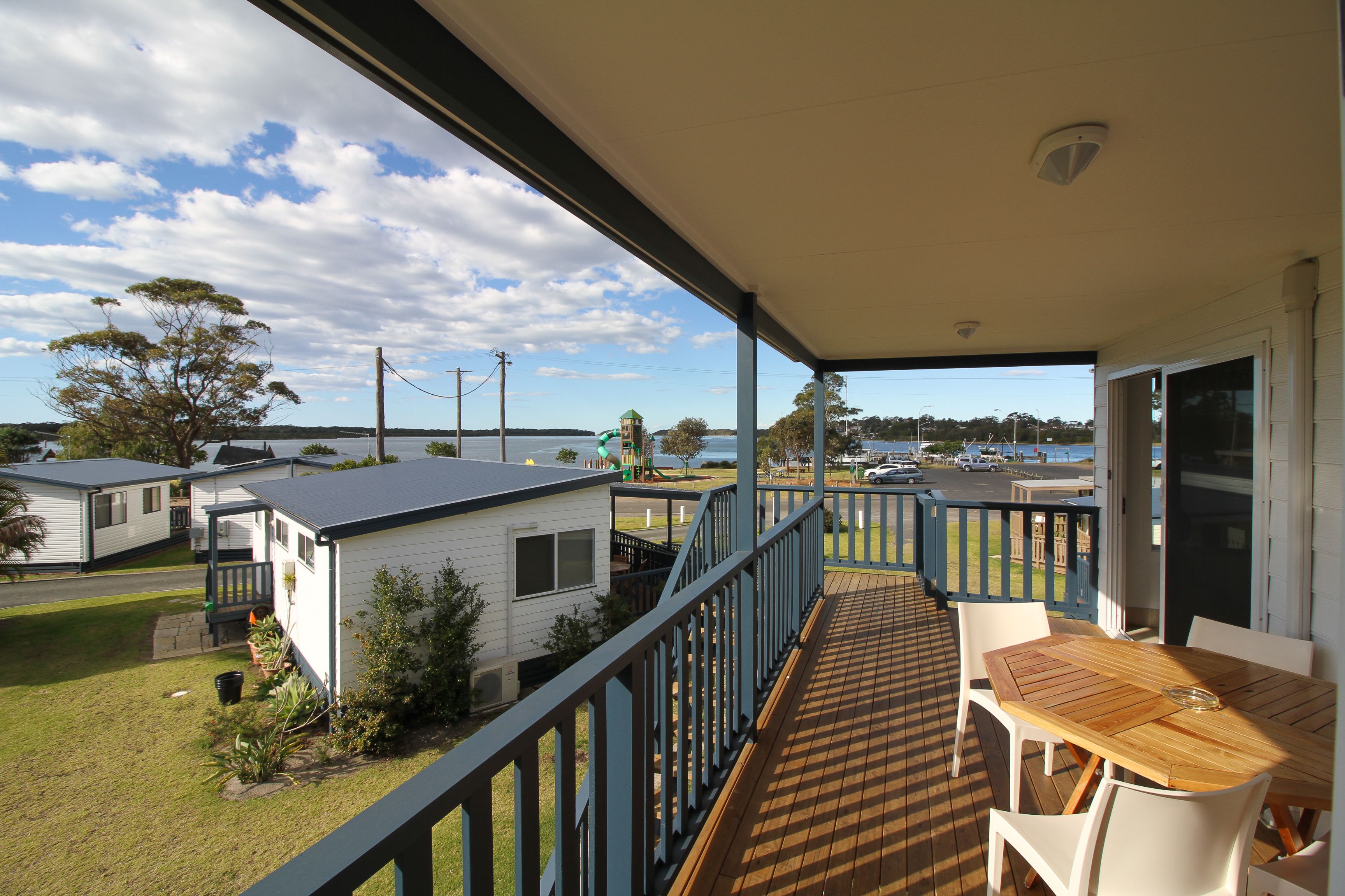 Un balcon avec une table et des chaises en bois, donnant sur un quartier résidentiel avec des maisons et une étendue d’eau.