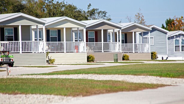 A row of mobile homes with front porches, a gravel driveway, and a grassy area.