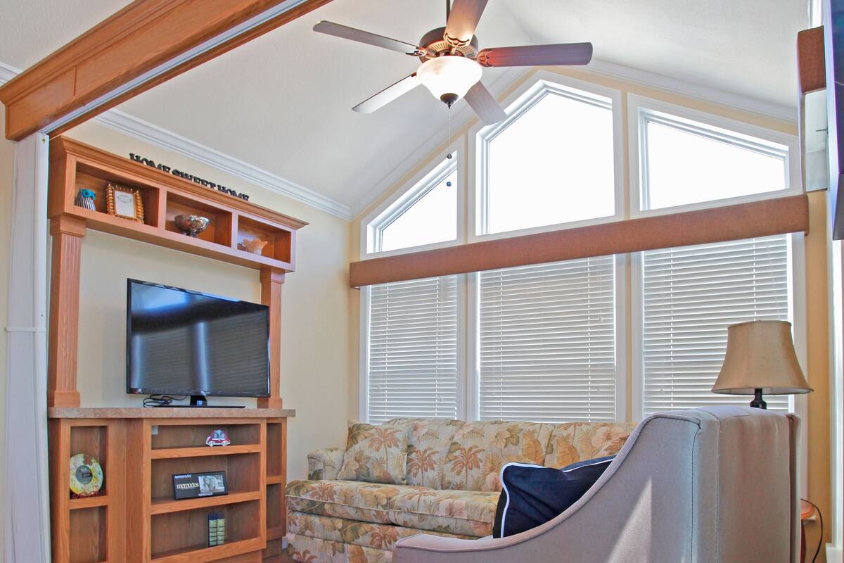 A living room with a television on a wooden stand, a sofa, a ceiling fan, and large windows with blinds.
