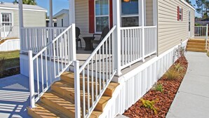 A single-story mobile home with a covered porch, wooden steps, and a small garden area.