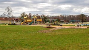 A playground with various play structures, including slides and climbing areas, surrounded by a grassy field and a building in the background. - Indian Lakes RV Campground (Sunman)