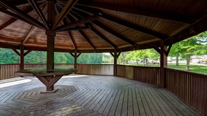 A covered wooden deck with a central table, surrounded by a railing and trees in the background. - Indian Lakes RV Campground (Sunman)