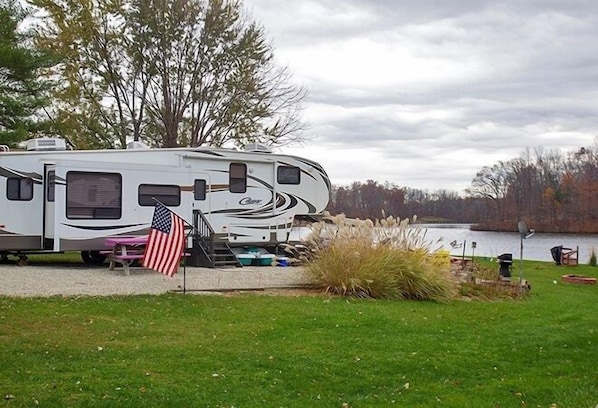 A white RV with a "Crown" logo parked near a lake, an American flag, and a picnic table.