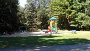 Une aire de jeux avec un toboggan et des structures d’escalade, entourée d’arbres et bordée d’une zone pavée.