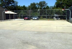 A parking area with a basketball hoop, a building with a sloped roof, and trees in the background. - Twin Mills RV Resort (Howe)