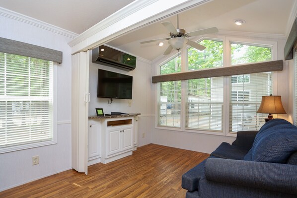A spacious living room with a ceiling fan, a built-in TV, and a large window with blinds.