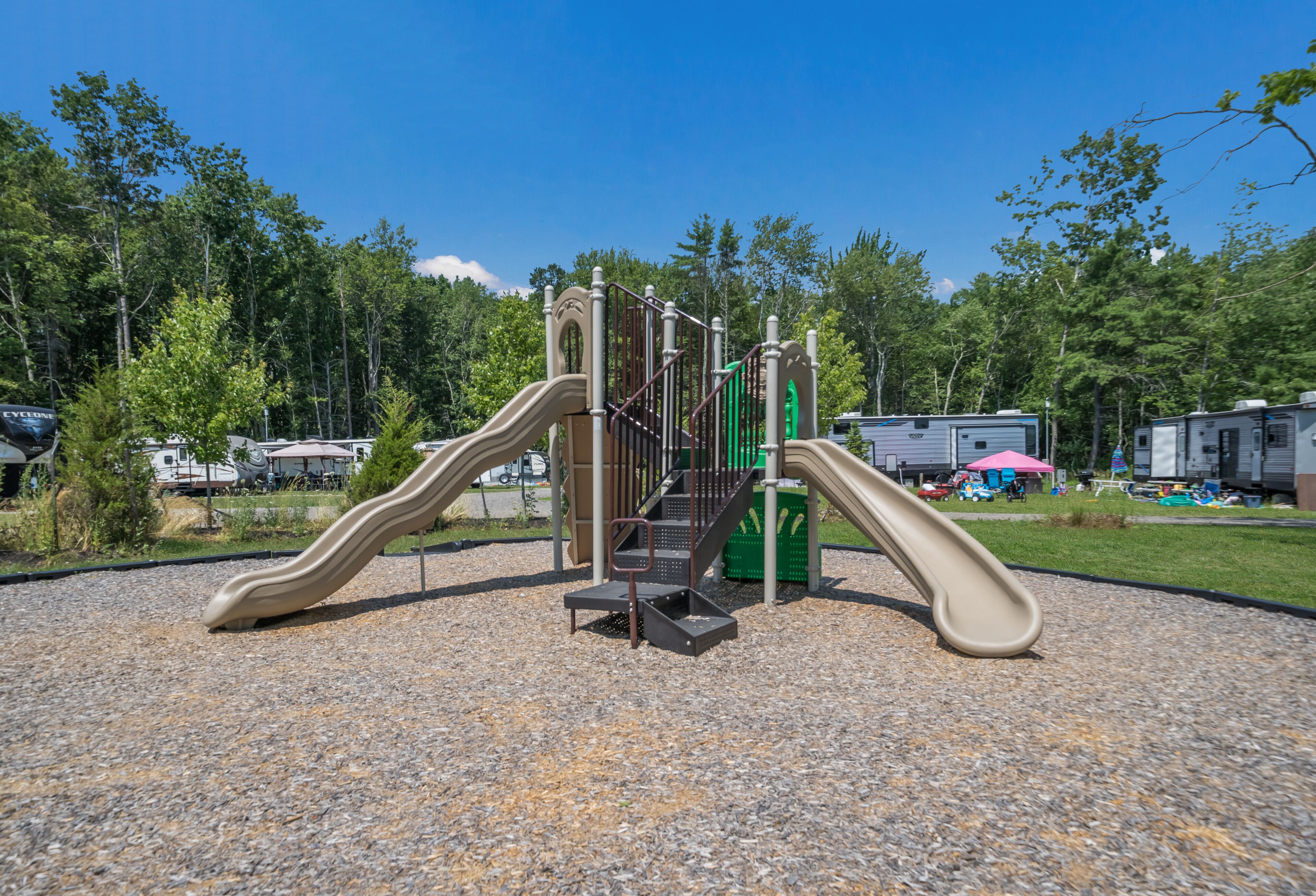 A playground with two slides and a climbing structure, surrounded by gravel and trees.