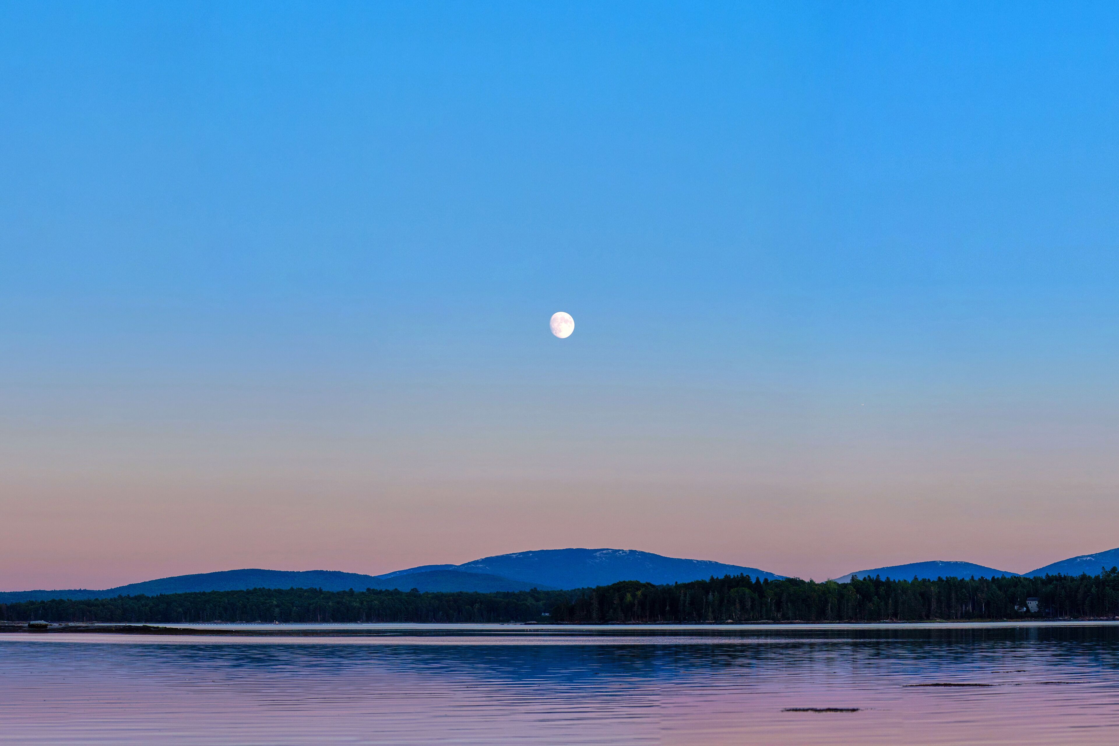 A calm lake at dusk with a visible moon and distant mountains.