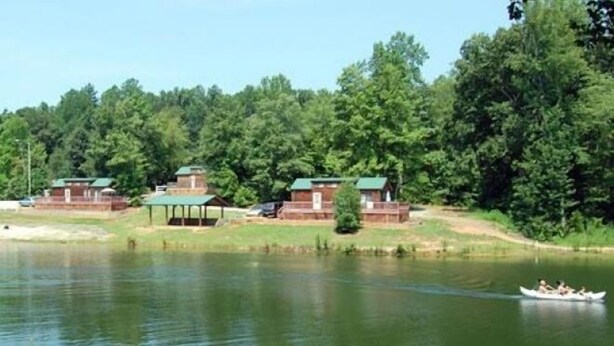 A lakeside property with cabins, a boat on the water, and dense forest in the background. - Forest Lake RV & Camping Resort (Advance)