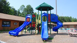 A playground with two blue slides, a green-roofed playhouse, and a building with a sign that reads "The Country Store." - Forest Lake RV & Camping Resort (Advance)