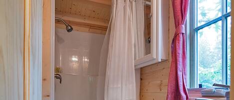 A wooden cabin bathroom with a shower, toilet, sink, and a window with a view of trees.