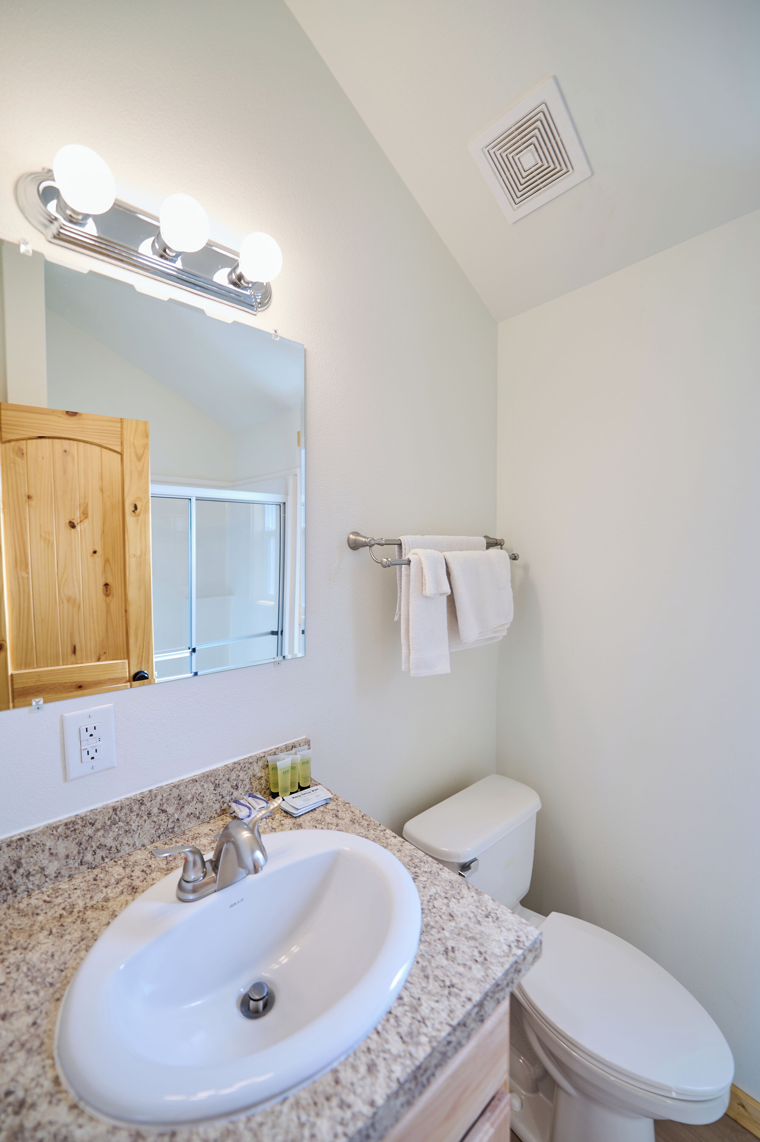 Een badkamer met een witte wastafel, meubel en toilet. Er is een spiegel met verlichting erboven, en een handdoekhouder met een handdoek eraan.