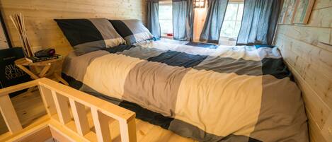 A wooden cabin bedroom with a bed, a nightstand, and a window with curtains.