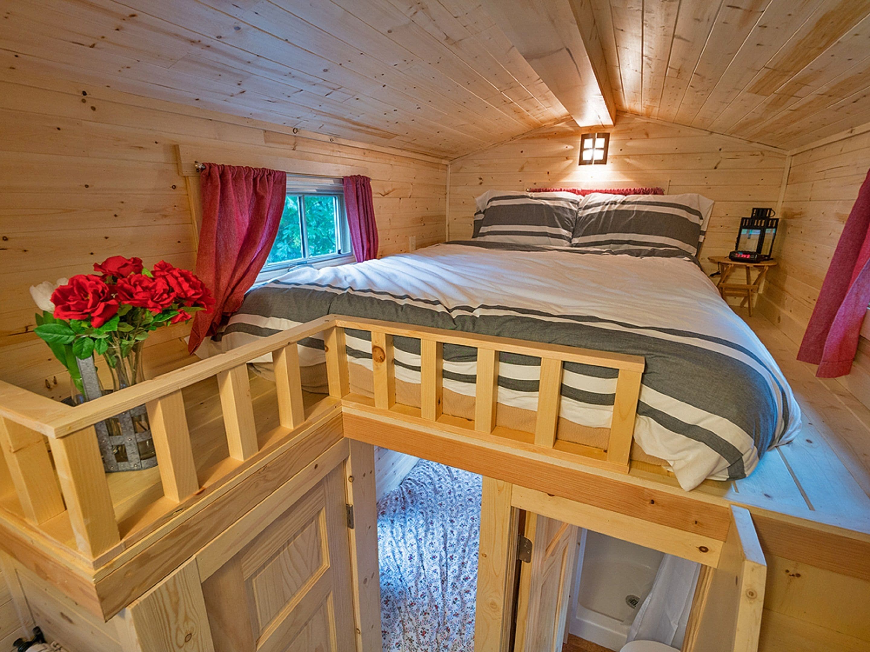 A wooden cabin bedroom with a bed, a window with curtains, a small table with a lamp, and a wooden staircase.