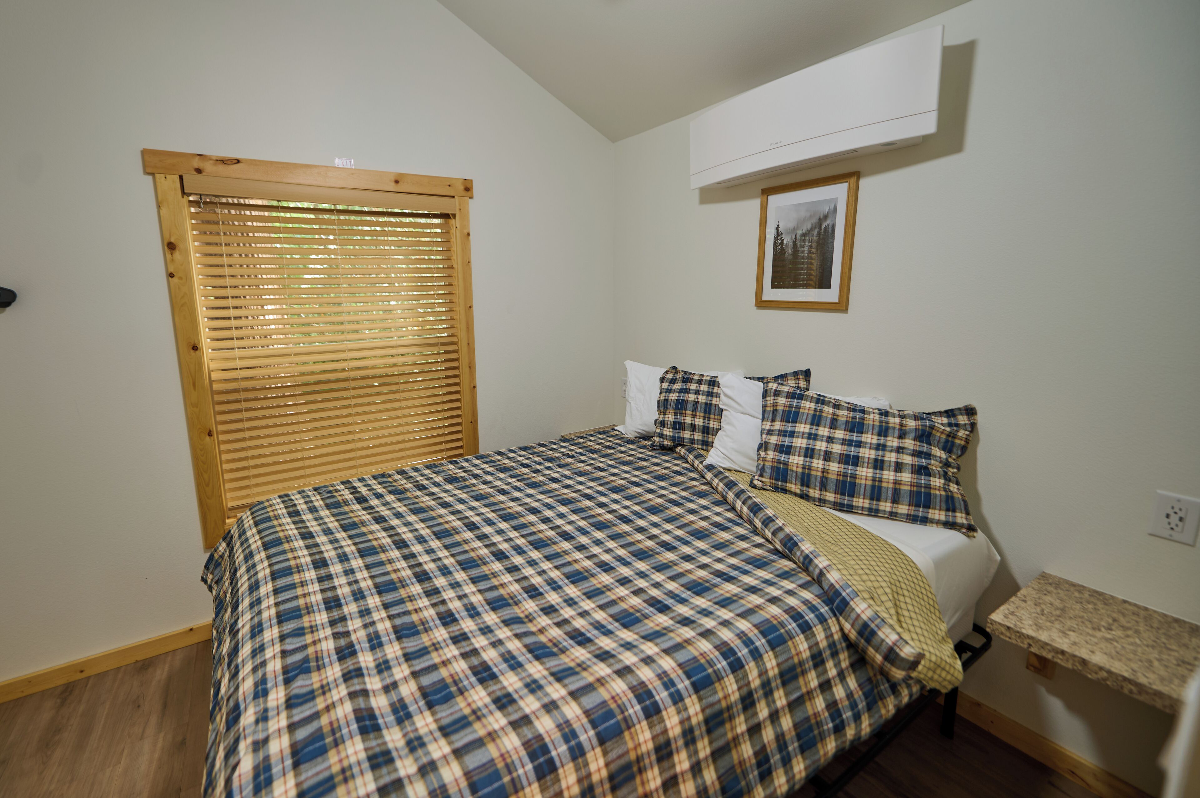 Ein Schlafzimmer mit kariertem Bettbezug, einem Holzjalousie, einem gerahmten Bild an der Wand und einem kleinen Regal.