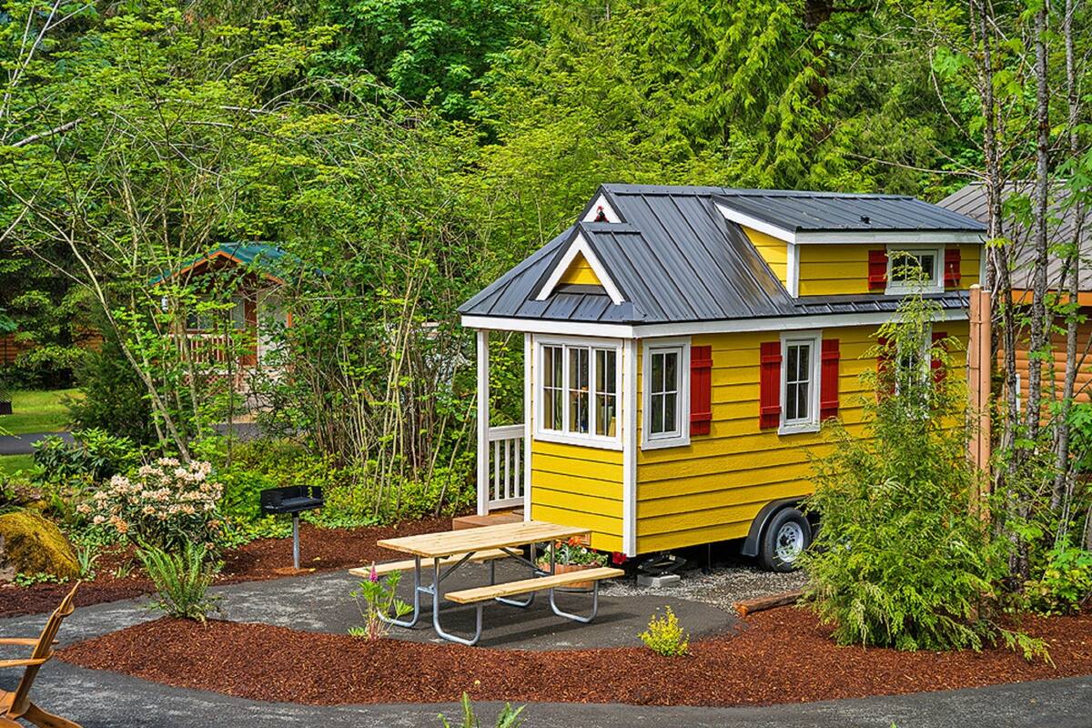 A small yellow house with a porch and a picnic table, surrounded by a garden and trees.