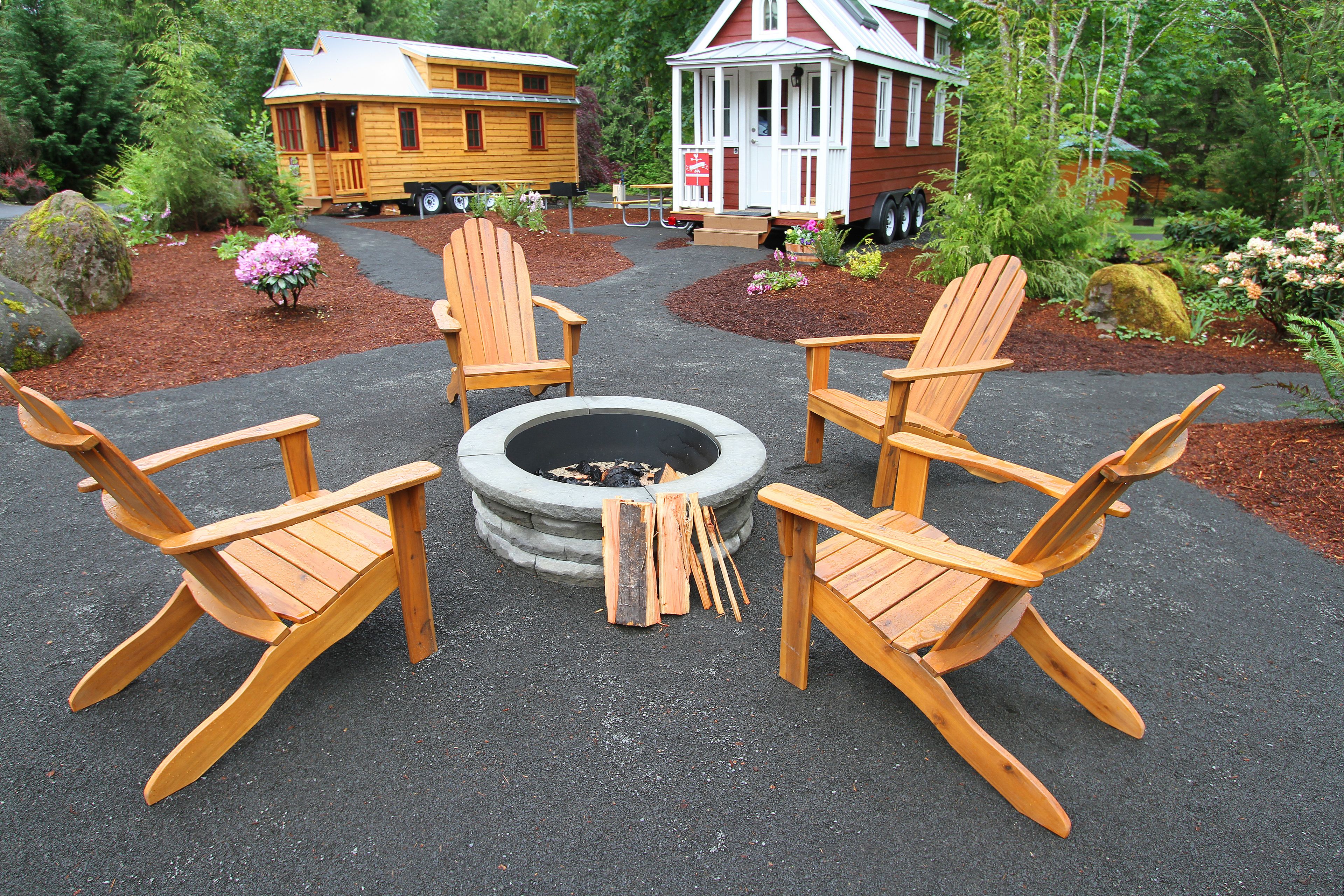 A fire pit with four wooden Adirondack chairs arranged around it, set in a landscaped area with mulch and plants.
