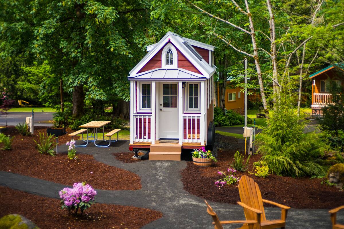 A small, single-story wooden structure with a white door and purple trim, surrounded by a garden with mulch paths, benches, and potted plants.