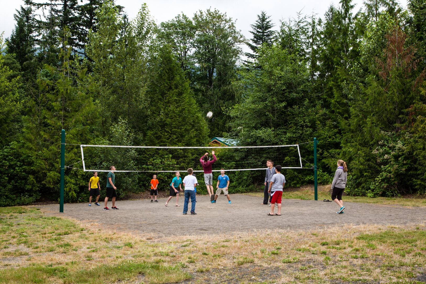 Eine Gruppe von Menschen spielt Beachvolleyball auf einem Sandplatz, umgeben von dichtem Wald.