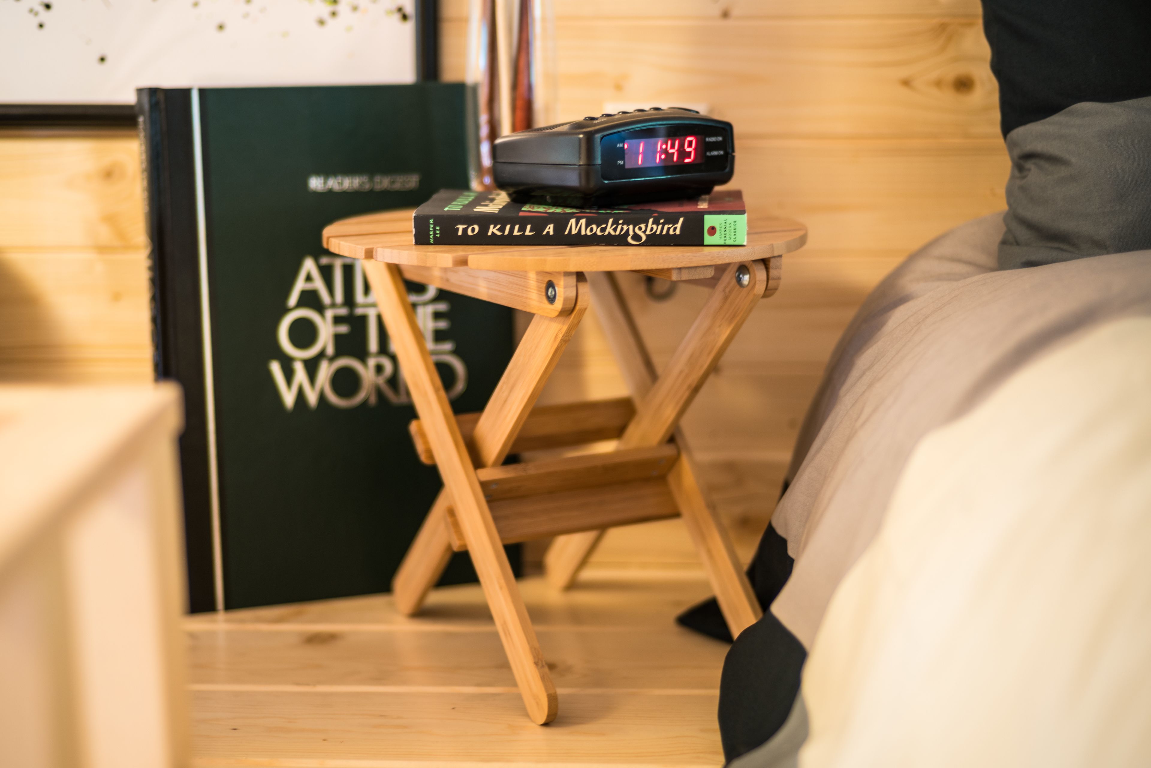 Ein Nachttisch mit einer digitalen Uhr, die 11:49 anzeigt, einem Buch mit dem Titel 'To Kill a Mockingbird' und einem Buch 'Atlas der Welt'.