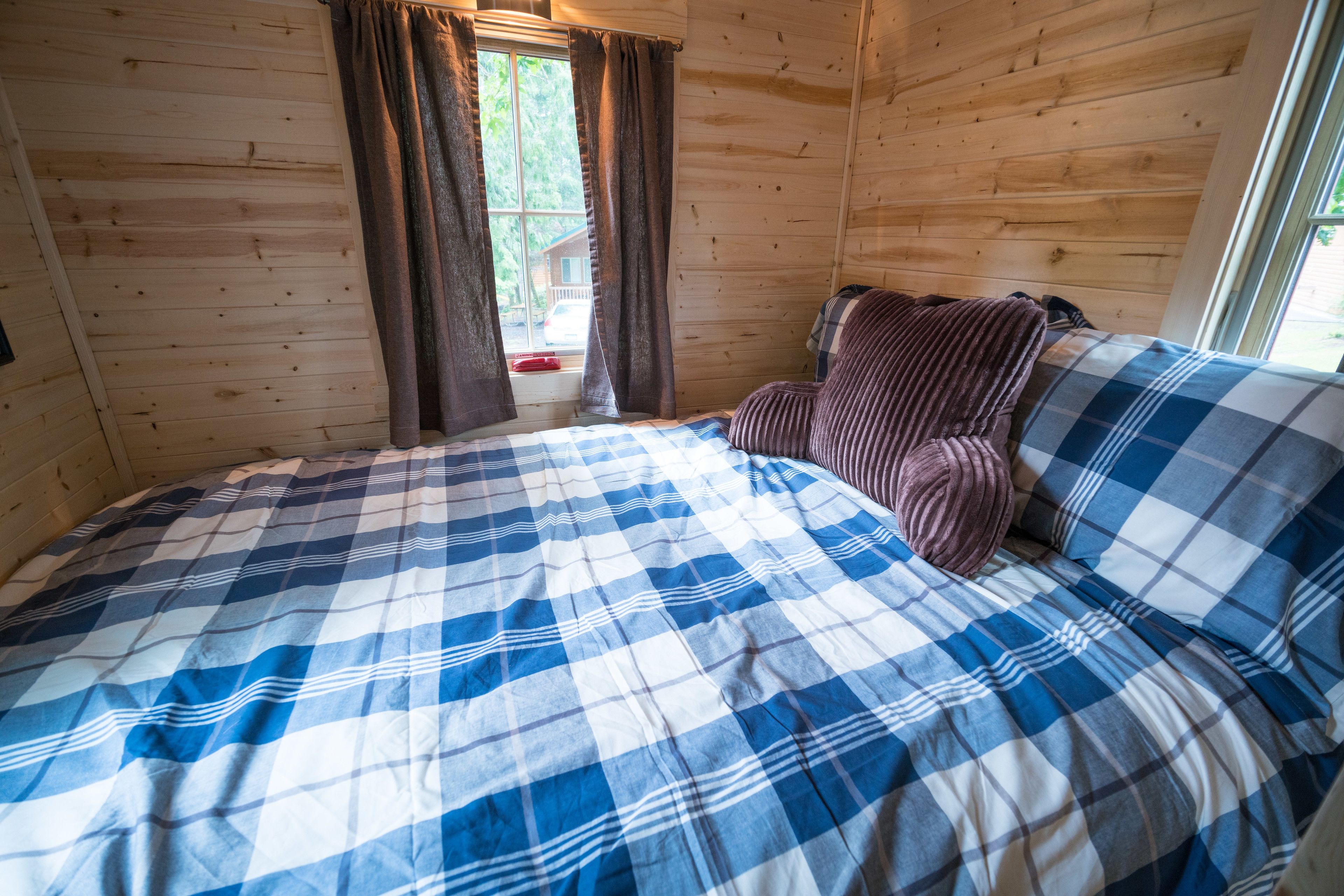 A wooden cabin bedroom with a bed, a window with curtains, and a view of a house outside.