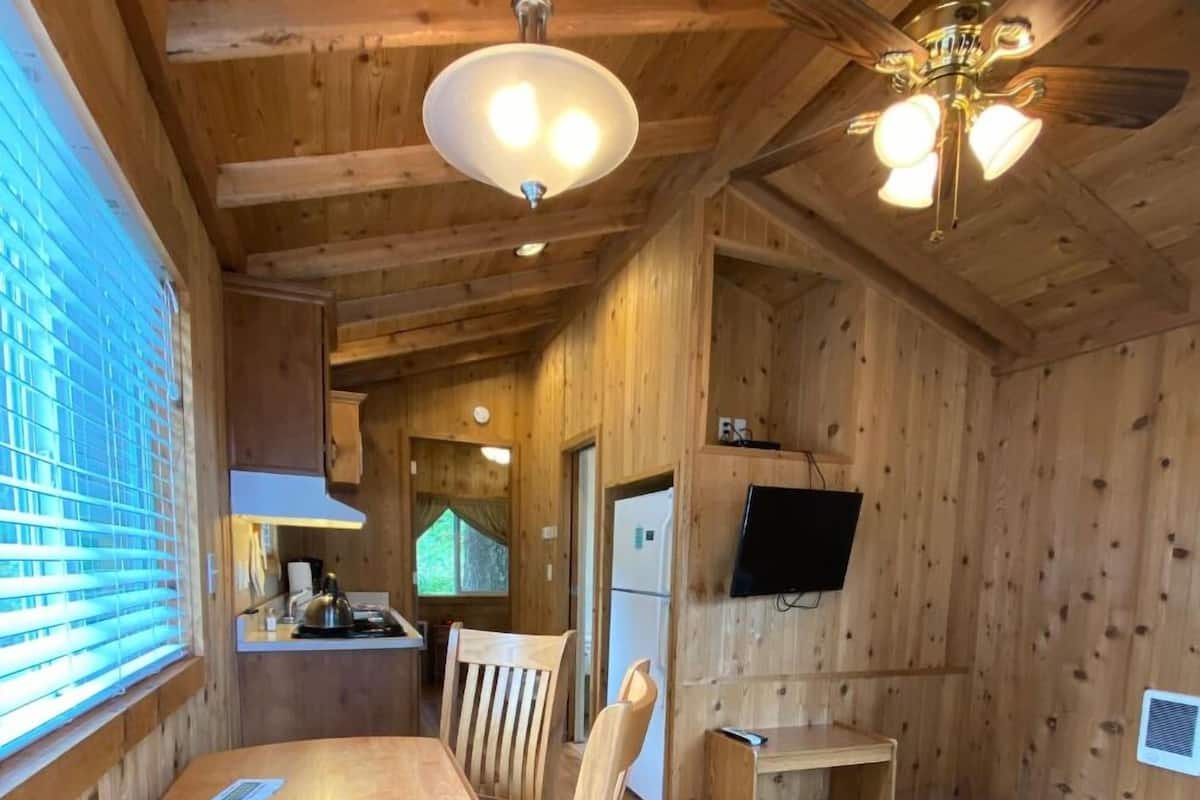 Interior de una cabaña de madera con una mesa de comedor, sillas, un ventilador de techo, un televisor montado en la pared y una pequeña zona de cocina.