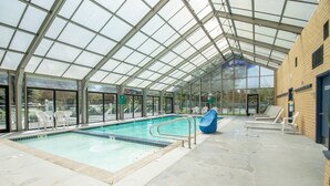 Una piscina interior con paredes de vidrio transparente, piso de baldosas y varias sillas de descanso.