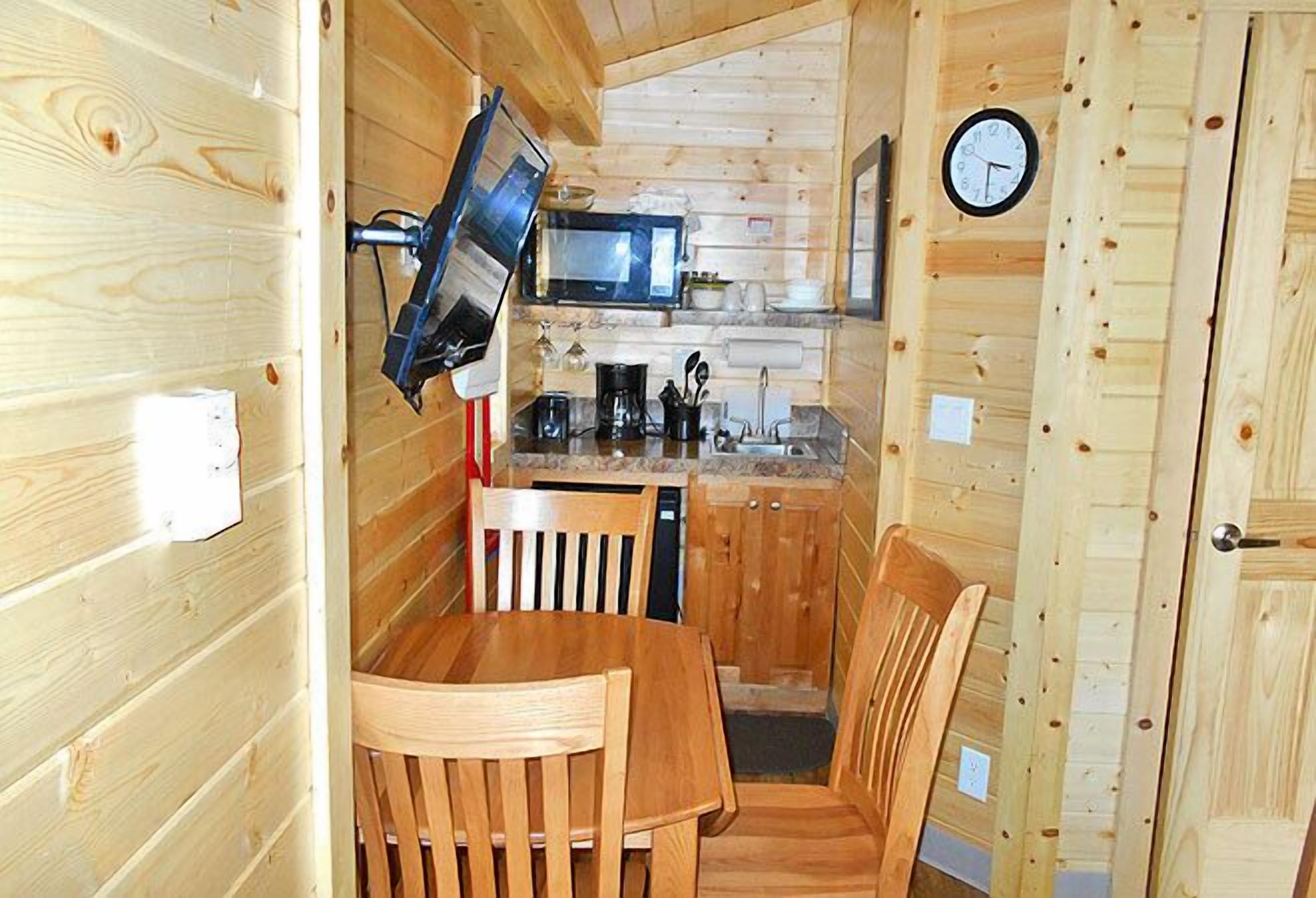 A compact kitchen with wooden walls, a dining table, chairs, a microwave, and a clock.