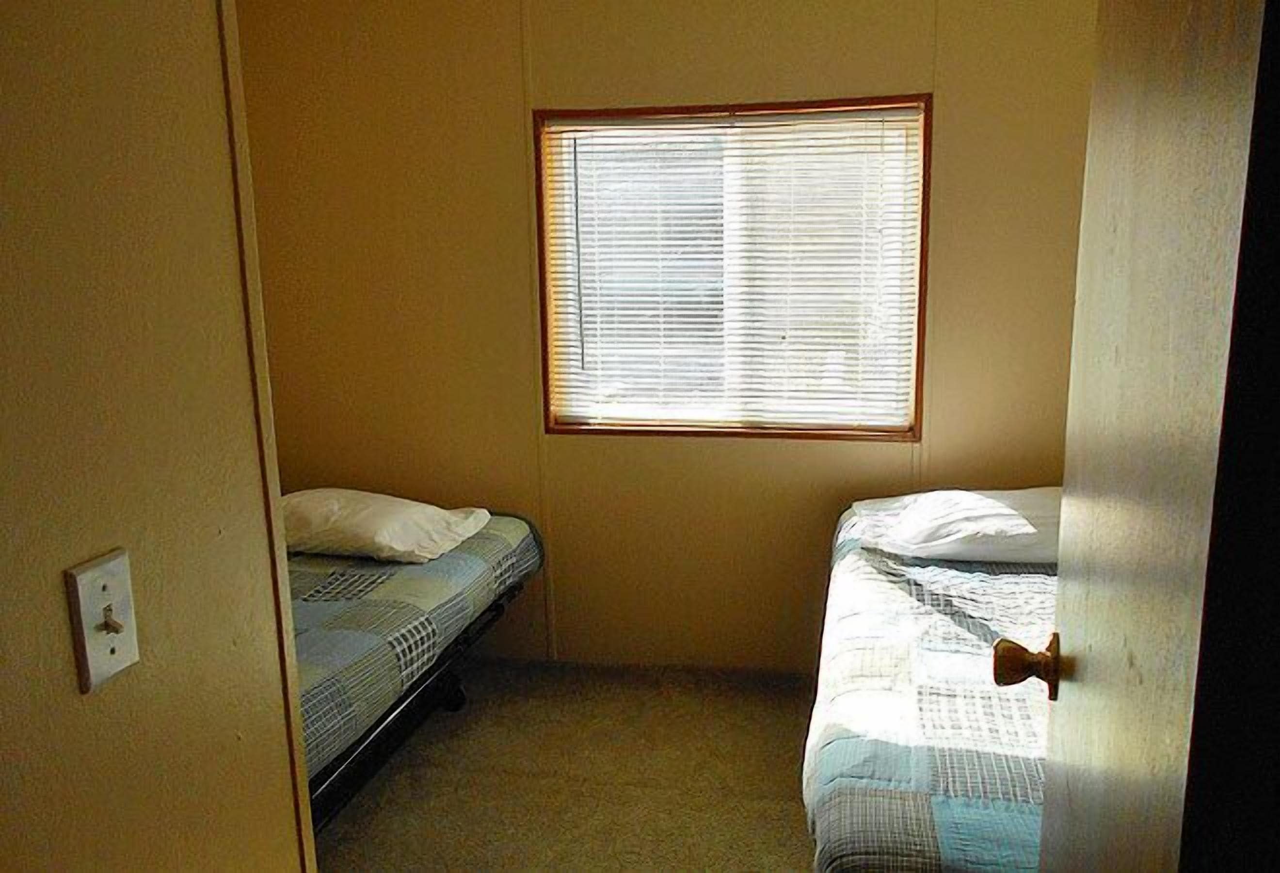 Um quarto pequeno e com pouca luz, com duas camas de solteiro, uma janela com persianas e uma porta.