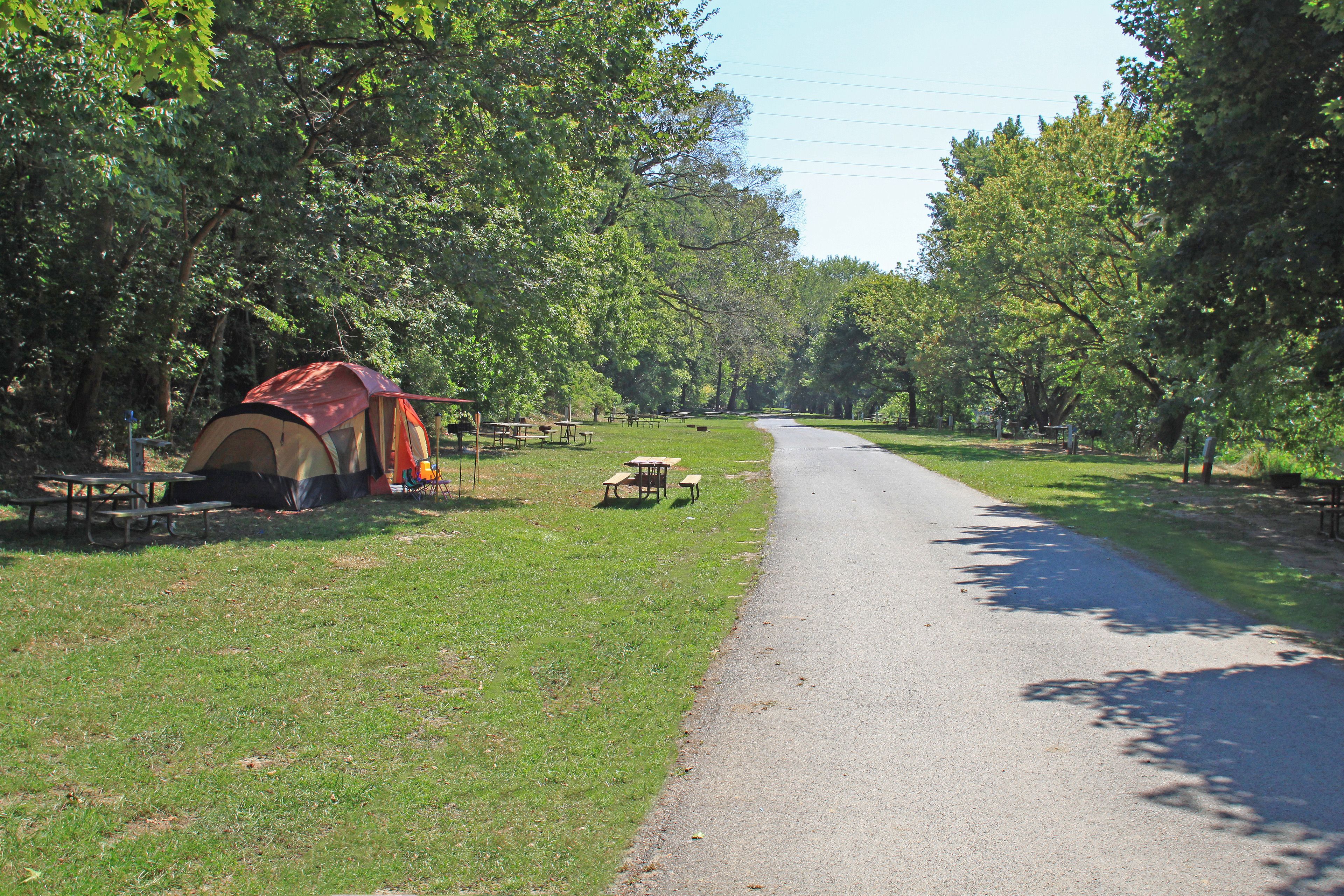 A campsite with a tent, picnic tables, and benches along a paved path surrounded by trees.