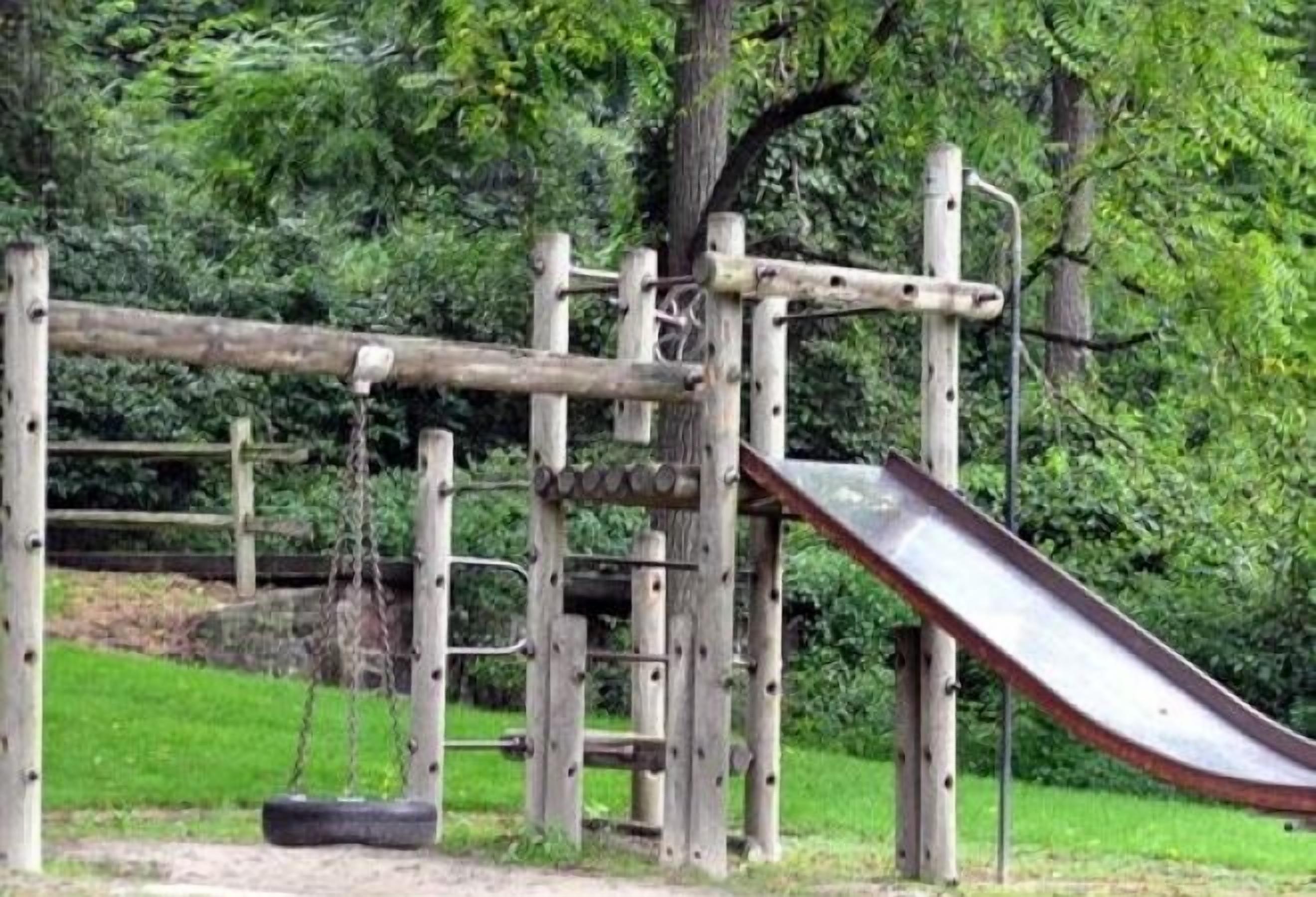A wooden playground structure with a slide and swings, surrounded by greenery.
