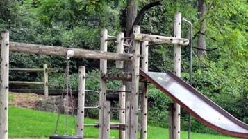 A wooden playground structure with a slide and swings, surrounded by greenery.