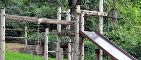 A wooden playground structure with a slide and swings, surrounded by greenery.
