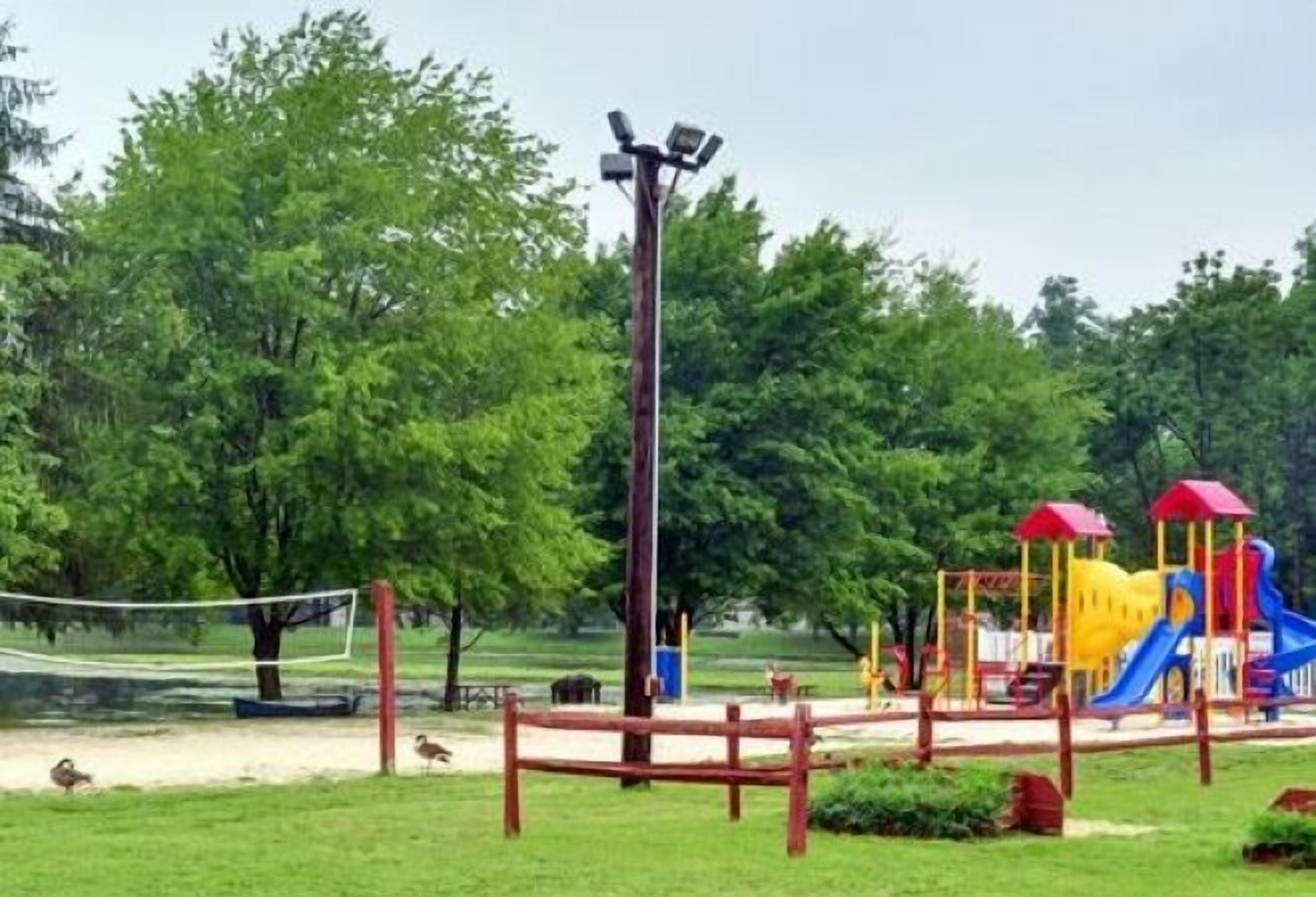 A playground with a slide and climbing structures, surrounded by trees and a volleyball net.