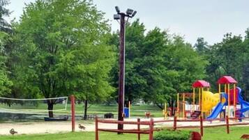 A playground with a slide and climbing structures, surrounded by trees and a volleyball net.