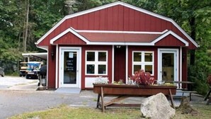 Un bâtiment rouge avec une porte et des fenêtres blanches, un banc en bois à l’extérieur et un grand rocher devant.