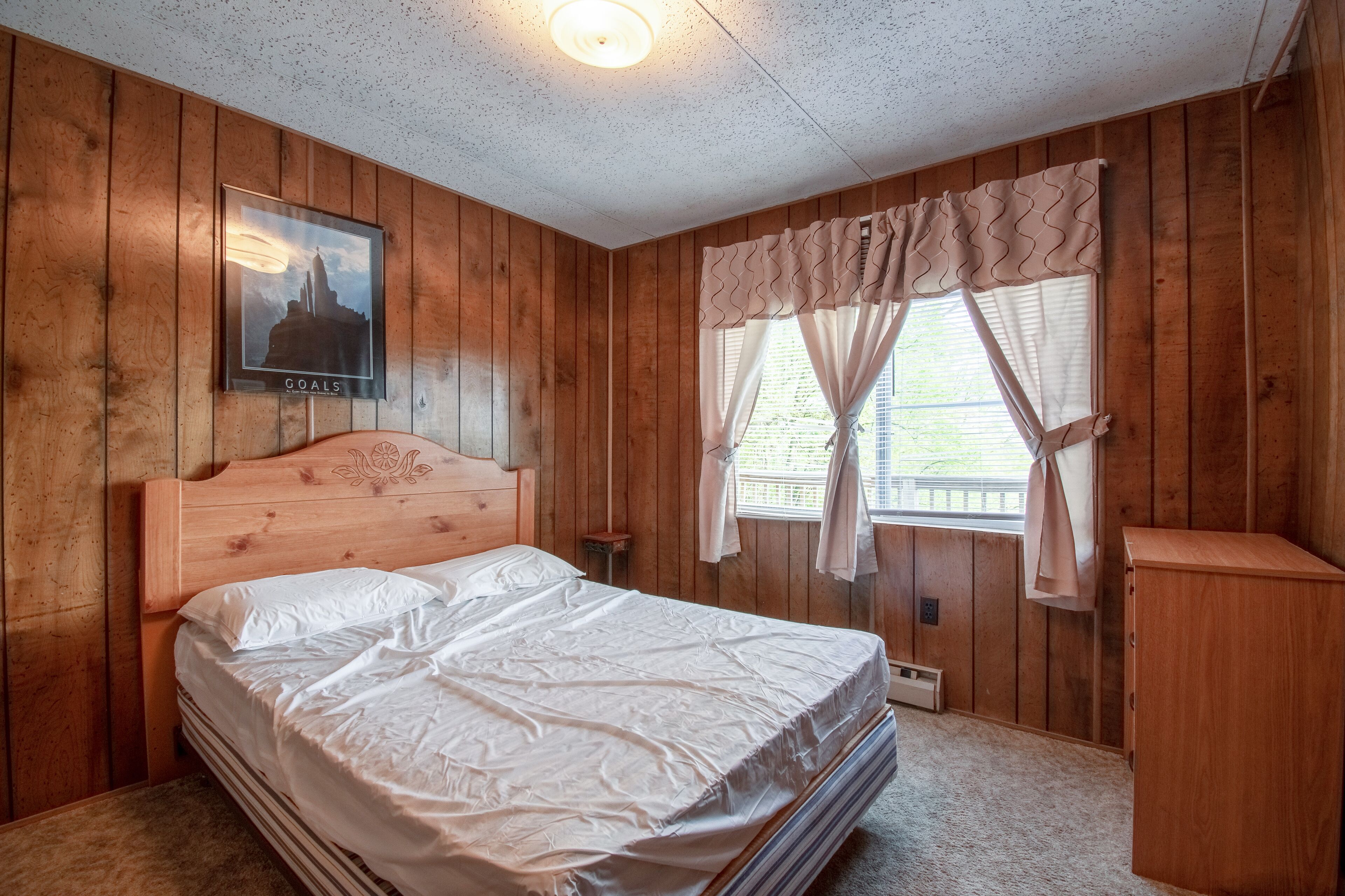 A wooden-paneled bedroom with a bed, a nightstand, a framed picture on the wall, and a window with curtains.