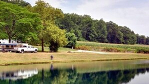 A white RV parked by a calm lake with trees and a field in the background. - Spring Gulch RV Campground (New Holland)