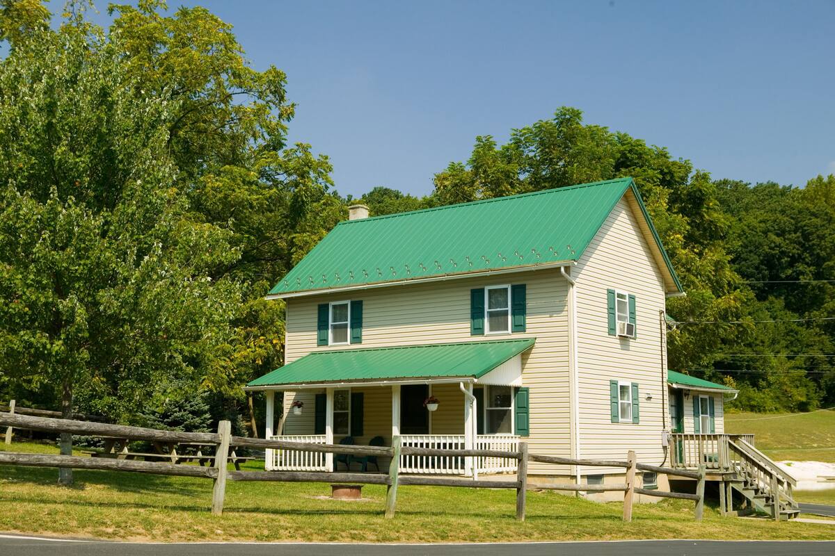 A two-story house with a green roof and white siding, featuring a porch with a railing and a small staircase leading to the front door.