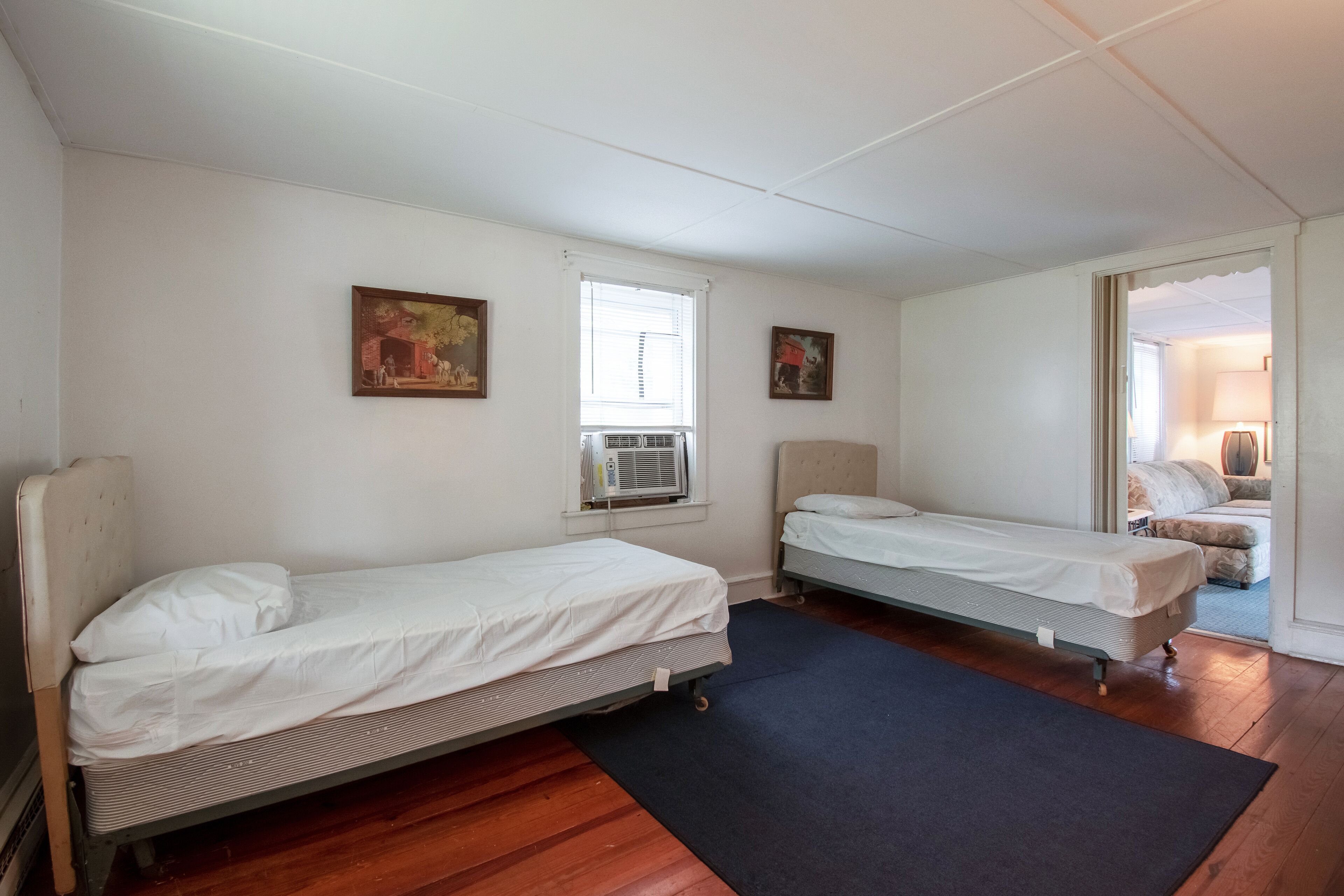 Une chambre avec deux lits simples, une fenêtre avec des stores, un appareil de climatisation et une porte donnant sur une autre chambre.