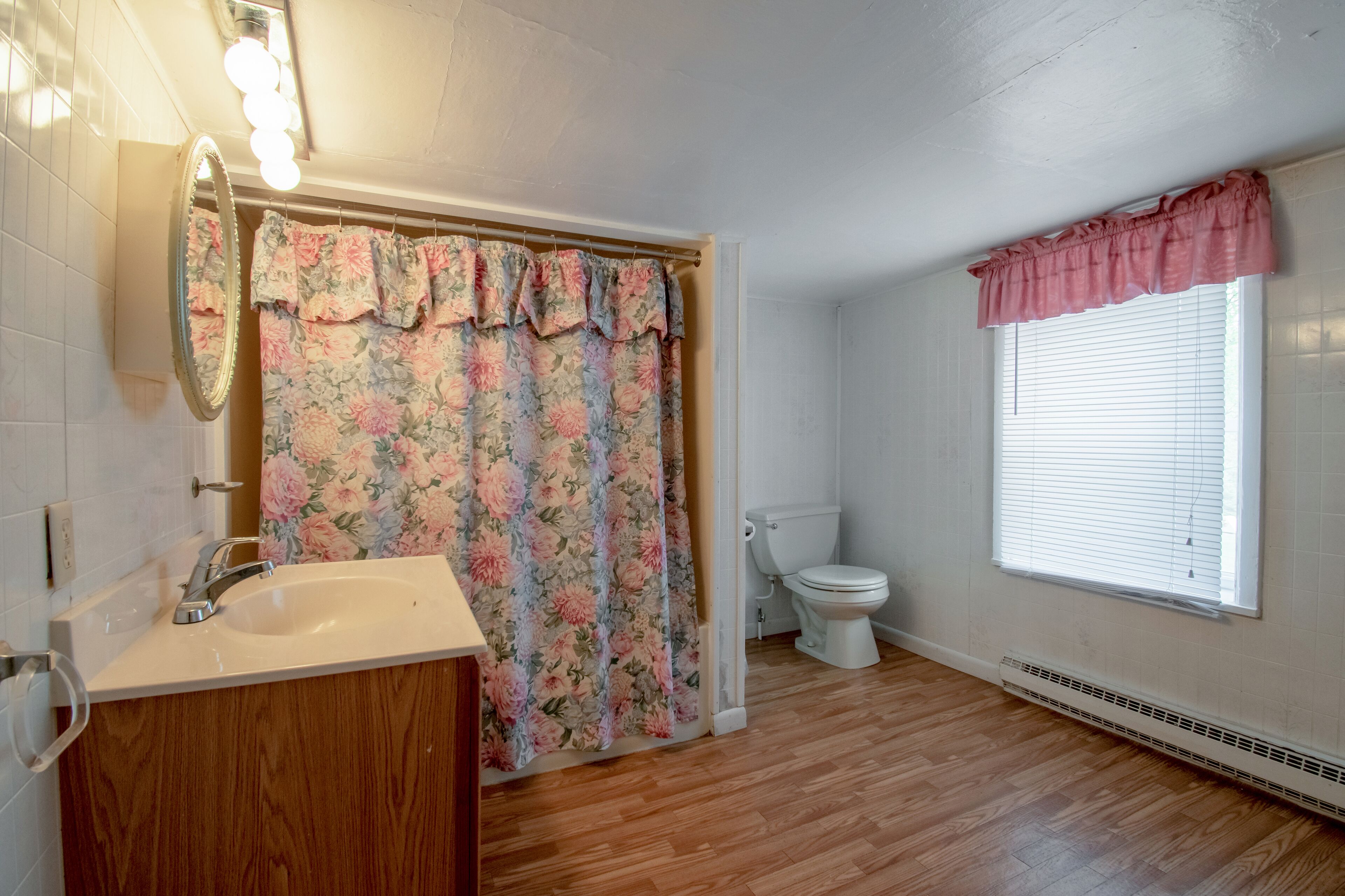 A bathroom with a wooden floor, a white sink, a mirror, a toilet, and a window with blinds.