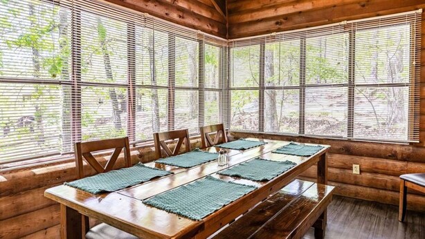 Une table à manger en bois, agrémentée de sets de table verts, entourée de chaises, dans une cabane dotée de grandes fenêtres et de murs en bois.